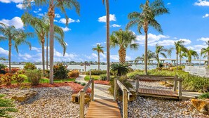 A pathway leading to a wooden bench overlooking a body of water, surrounded by palm trees and a bridge in the background.