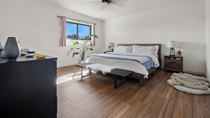 Un dormitorio con cama, mesitas de noche, un escritorio, una planta y una ventana con cortinas.