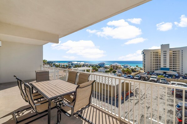 A balcony with a table and chairs, overlooking a parking area and a view of the ocean and buildings.