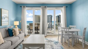 A living room with a sofa, coffee table, and dining area with chairs. There is a balcony with a view of buildings and the ocean.