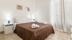 A hotel room with a bed, a nightstand with a lamp, two wall-mounted artworks, and a window with sheer curtains. - GuestHost - Cozy 87 sqm apartment, for 6 people, located on the first floor of a building (NO lift). The accommodation is located in the heart of the historic Ballarò market (in a restricted traffic zone), an area where it is possible to savor the true  (Palermo)