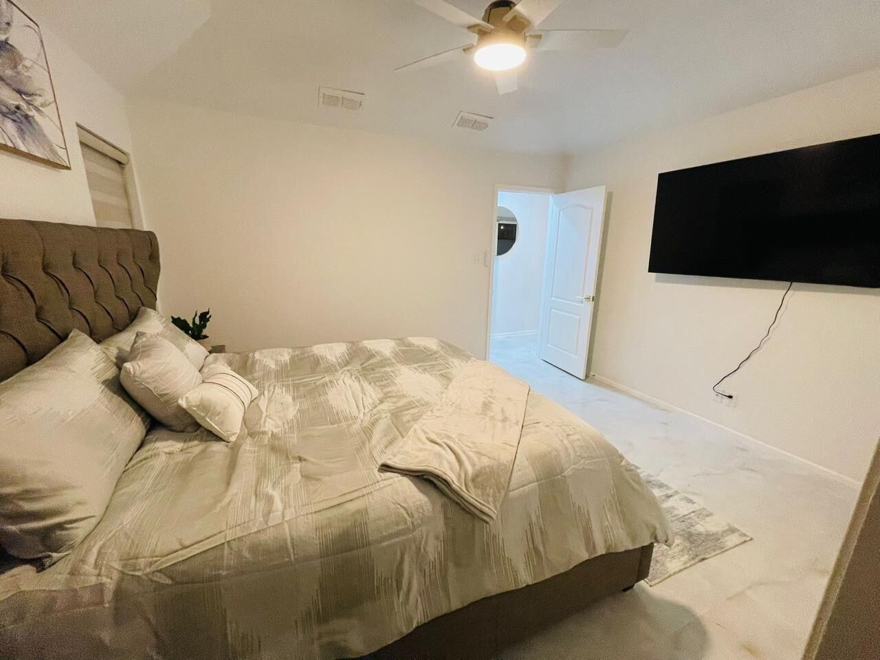 A bedroom with a bed, a ceiling fan, a TV mounted on the wall, and a door leading to another room.