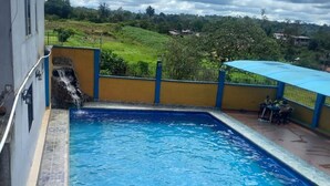 A rectangular swimming pool with a stone border, a small waterfall feature, and a view of a green landscape in the background. - Hotel Cristal Azul (Pedro Vicente Maldonado)