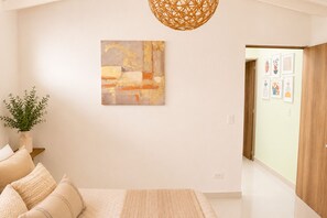 Ein modernes Schlafzimmer mit einem Bett, einem an der Wand befestigten abstrakten Gemälde, einer hängenden gewebten Leuchte und einem Flur, der zu einer Tür mit gerahmten Kunstwerken führt.