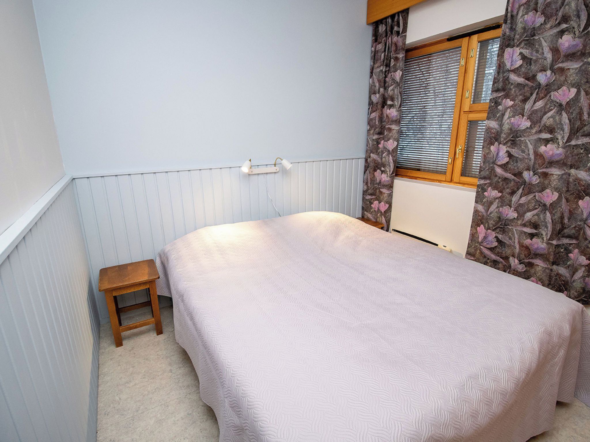A bedroom with a bed, a small wooden stool, a radiator, and a window with floral curtains.