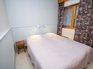 Ein Schlafzimmer mit einem Bett, einem kleinen Holzschemel, einem Heizkörper und einem Fenster mit blumigem Vorhang.
