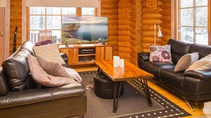 Een blokhut interieur met een houten tv-meubel, leren banken, een bijzettafeltje en een groot raam met uitzicht op een meer.