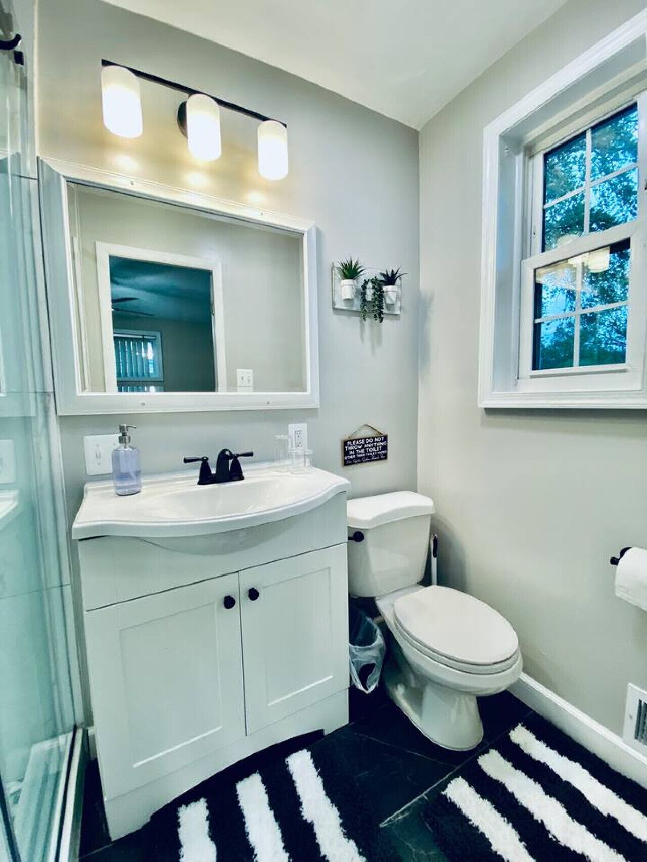 Een badkamer met een wit wastafelmeubel, een spiegel, een toilet en een raam met uitzicht op bomen.