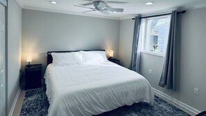 Ein Schlafzimmer mit einem Bett, Deckenventilator, zwei Nachttischlampen, einem Fenster mit Vorhängen und einem Nachttisch.