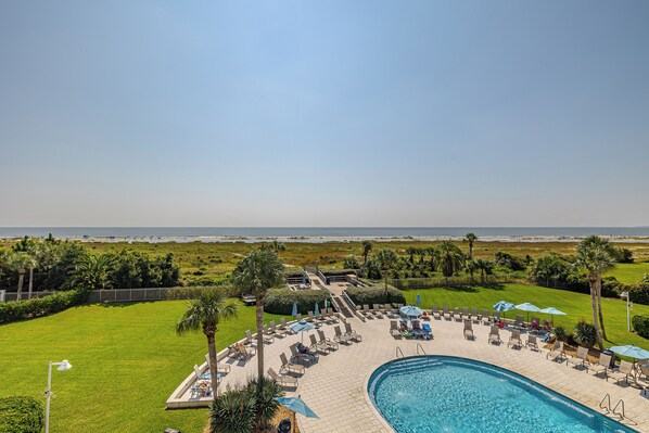 A pool area with lounge chairs and umbrellas, a clear view of the ocean, and a well-maintained lawn.
