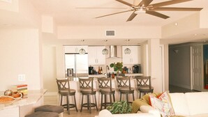 Cozinha moderna com armários brancos, uma ilha central e banquetas. Há um ventilador de teto sobre a cozinha.