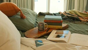 Quarto com cama, janela com cortinas, mesa de cabeceira com livros e uma caneca, e vista para a praia.