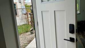 Eine weiße Tür mit Fenster und Klingel, die zu einem überdachten Außenbereich mit einem Betonweg und einem kleinen Garten führt.