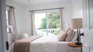 Ein Schlafzimmer mit einem Bett, einem Nachttisch mit Lampe, einem Fenster mit Vorhängen und Blick auf Grünfläche.