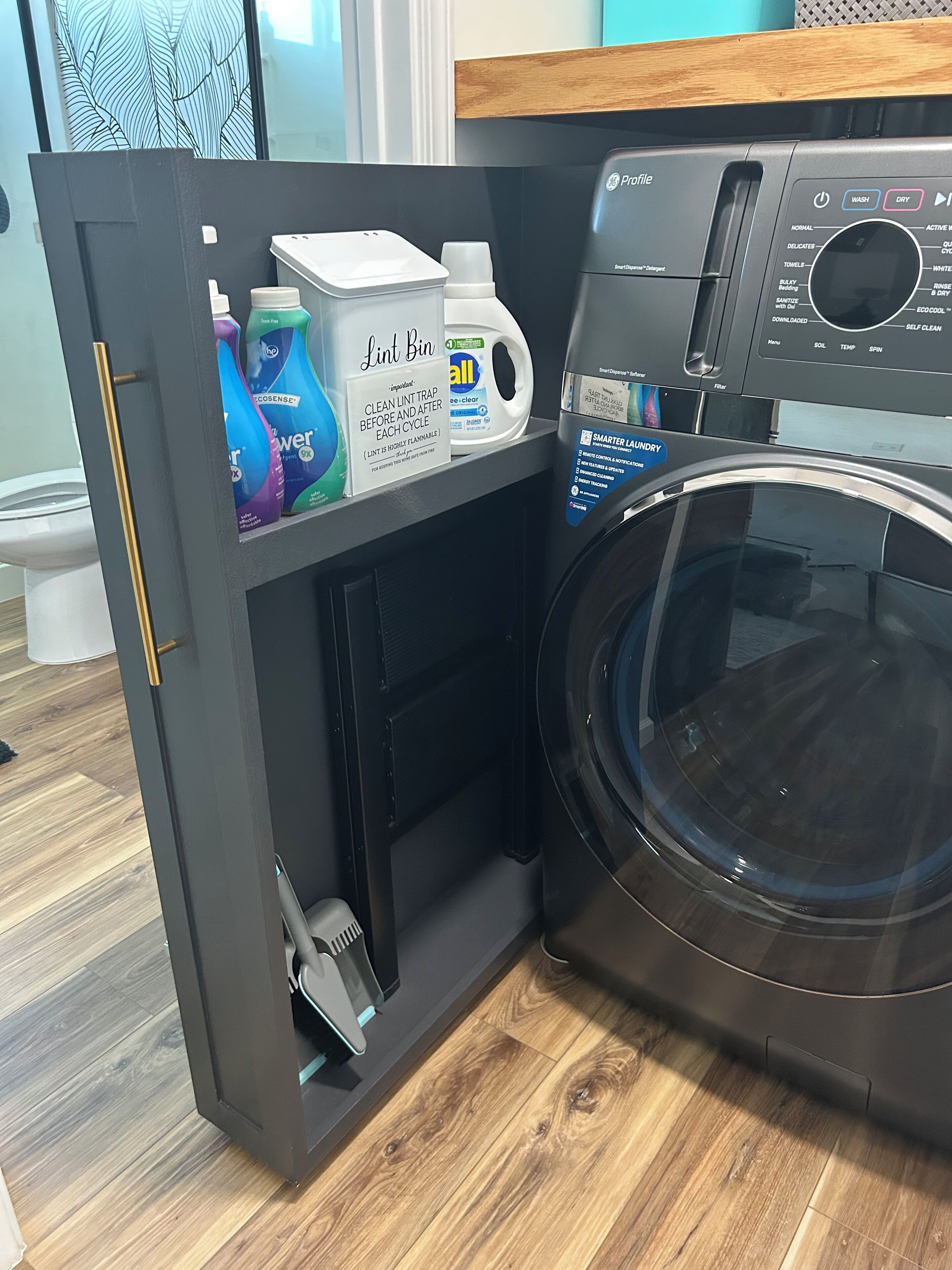 Een compacte was- en droogcombinatie met een plintplank, een waterfles en schoonmaakmiddelen.