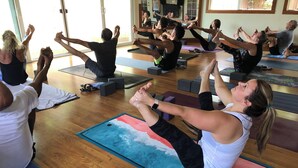 Eine Gruppe von Menschen praktiziert Yoga auf Matten in einem Raum mit Holzböden und großen Fenstern.