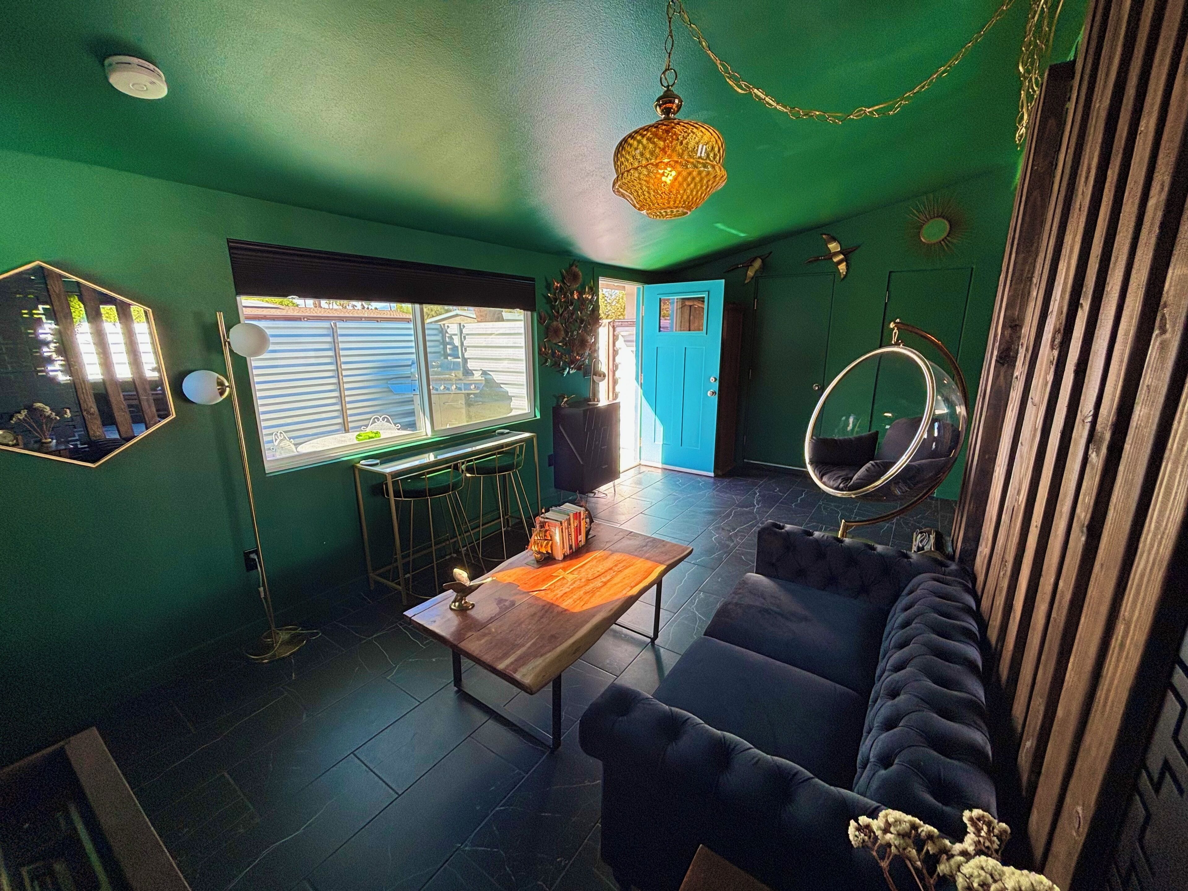 Una stanza con una parete verde, una porta blu, una poltrona sospesa, uno specchio rotondo e un tavolino da caffè in legno.