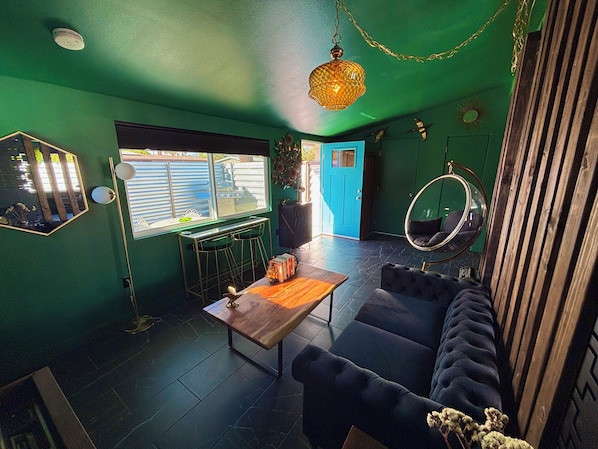 A room with a green wall, a blue door, a hanging chair, a round mirror, and a wooden coffee table.