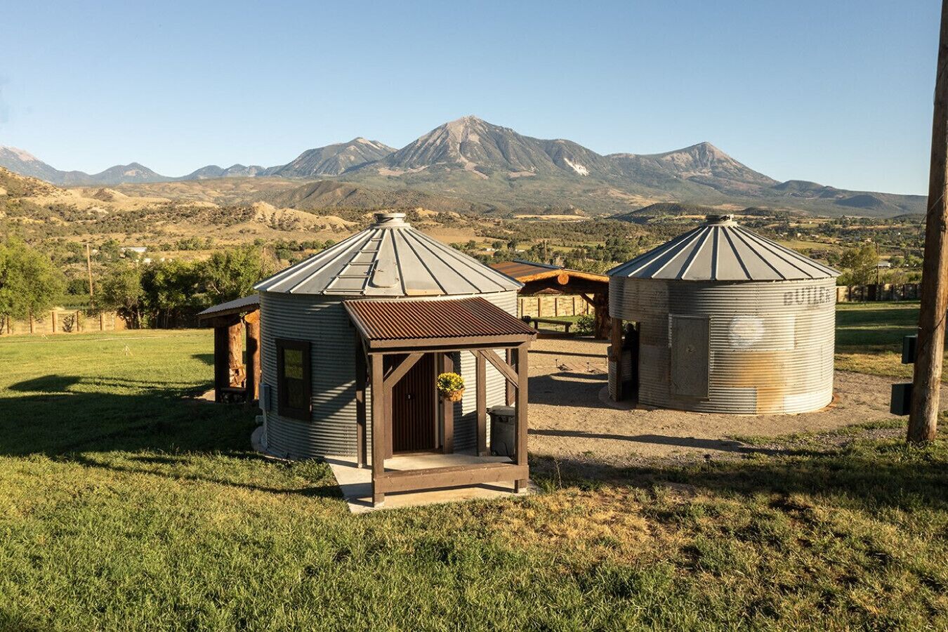Un granero rústico de estructura de madera y un silo en un campo cubierto de pasto, con montañas al fondo.