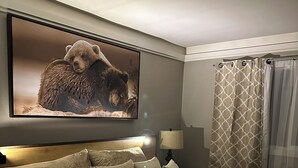 Una cama bien hecha con una colcha estampada, un cabecero con una lámpara y un cuadro de un oso colgado en la pared, además de una cortina estampada.