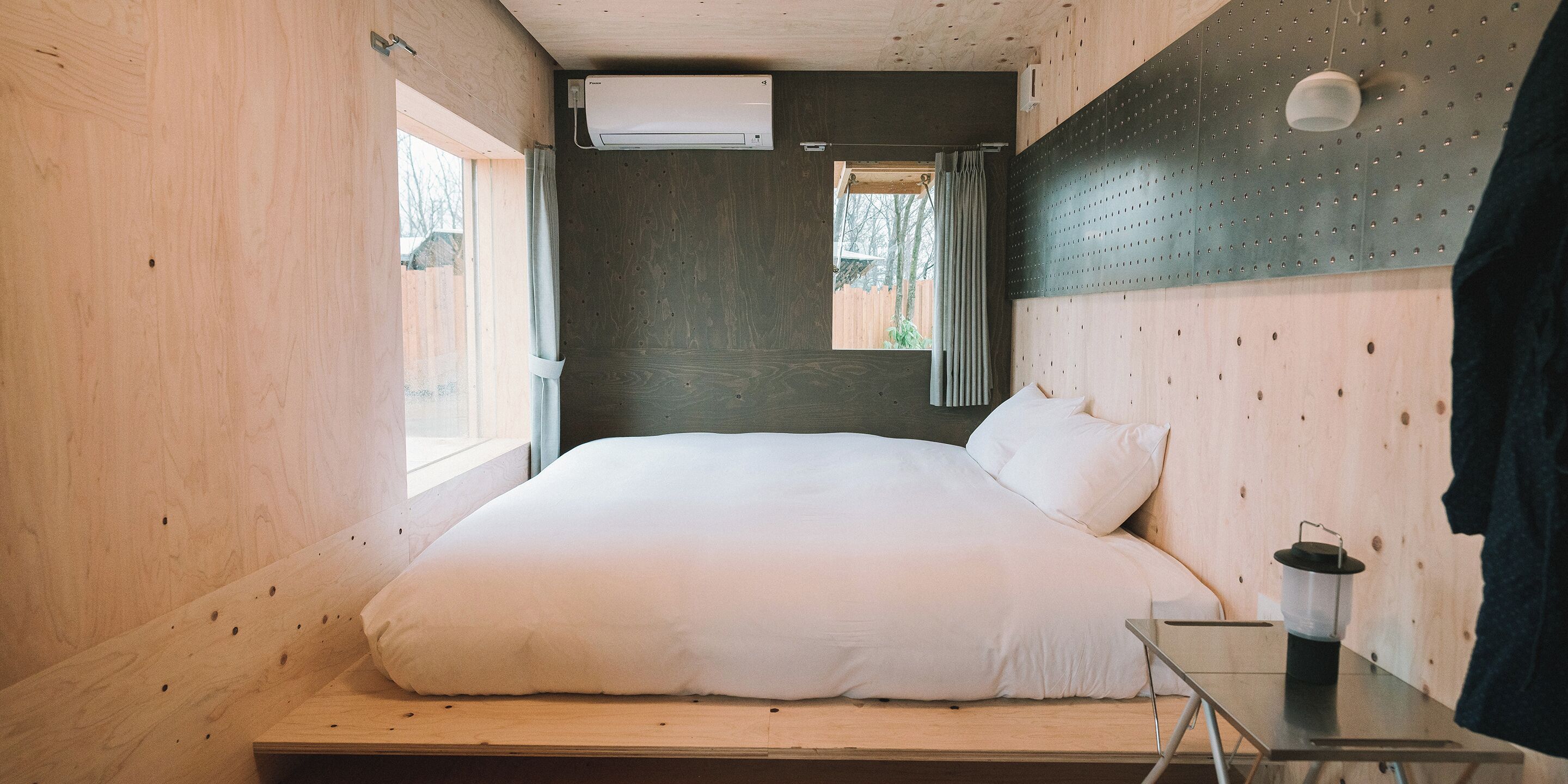 Une chambre à coucher de taille modeste, dotée d’une tête de lit en bois, d’un lit avec une literie blanche, d’une petite table de chevet et d’une fenêtre avec des rideaux.