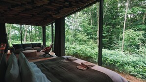 Une chambre moderne dotée d’un grand lit, d’un coin salon avec un fauteuil et un canapé, et offrant une vue sur une forêt dense grâce à de grandes fenêtres.