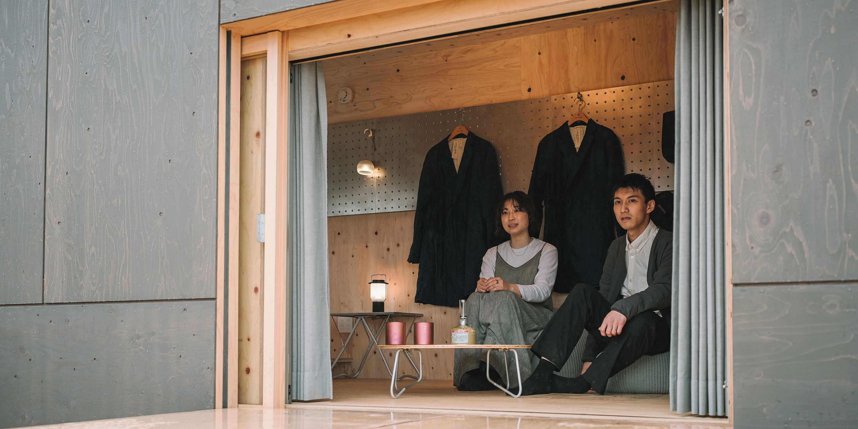 A room with wooden walls, a small table with a lamp, and three hangers with clothes.