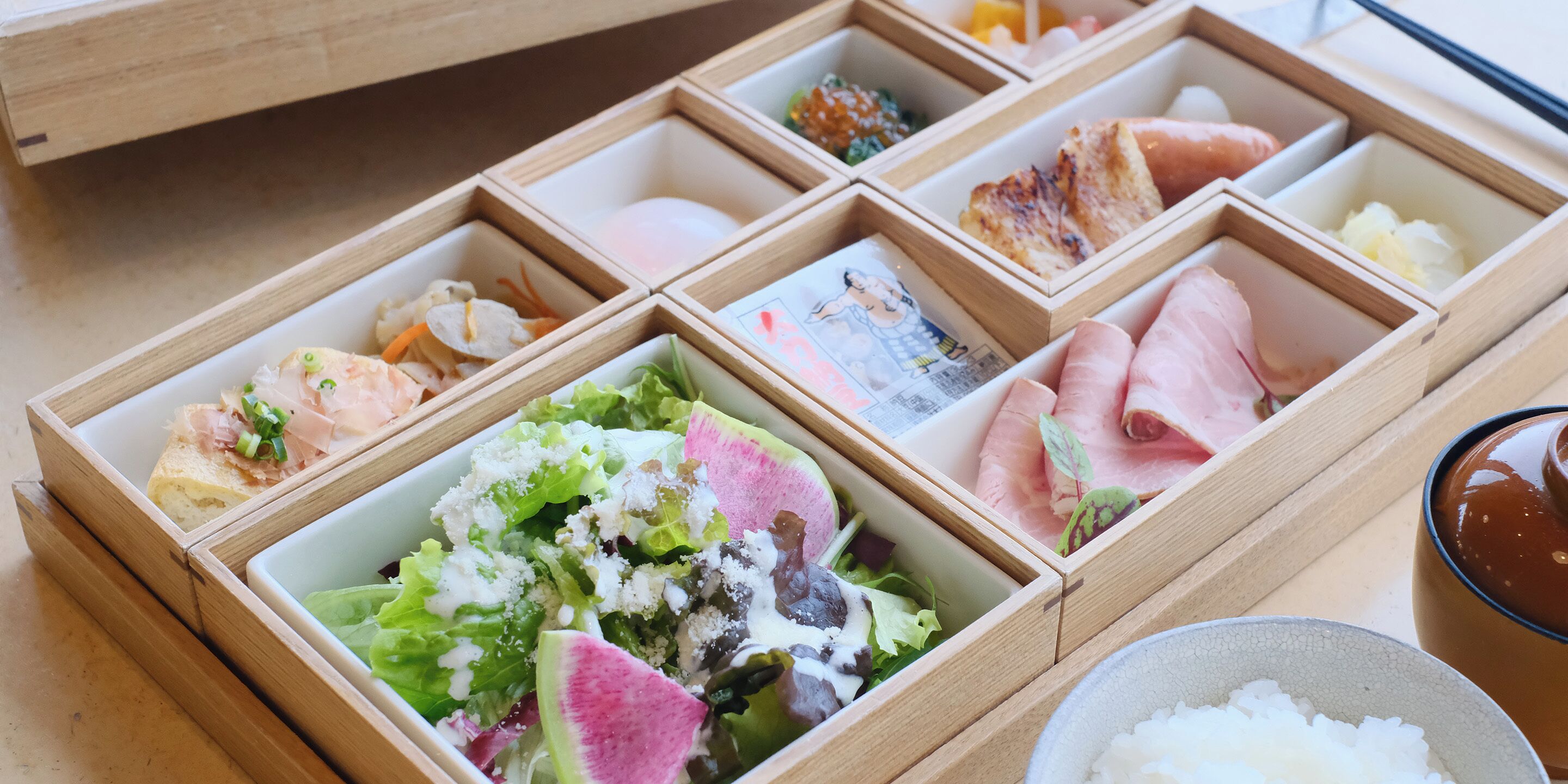 A wooden tray with compartments containing various food items and a bowl of rice.