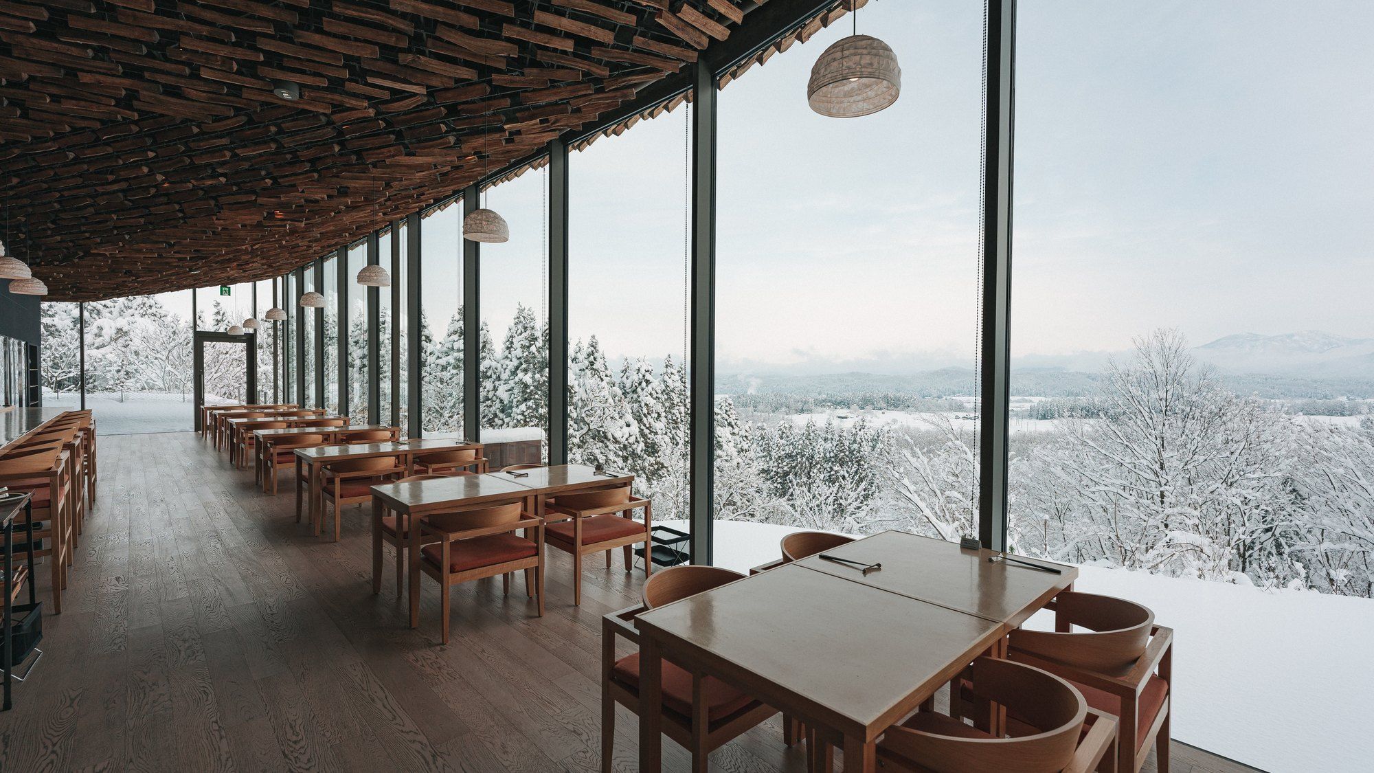 A restaurant with wooden tables and chairs, large windows, and a view of snow-covered trees.