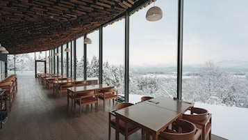 Un restaurante con mesas y sillas de madera, ventanas amplias y vistas a árboles cubiertos de nieve.
