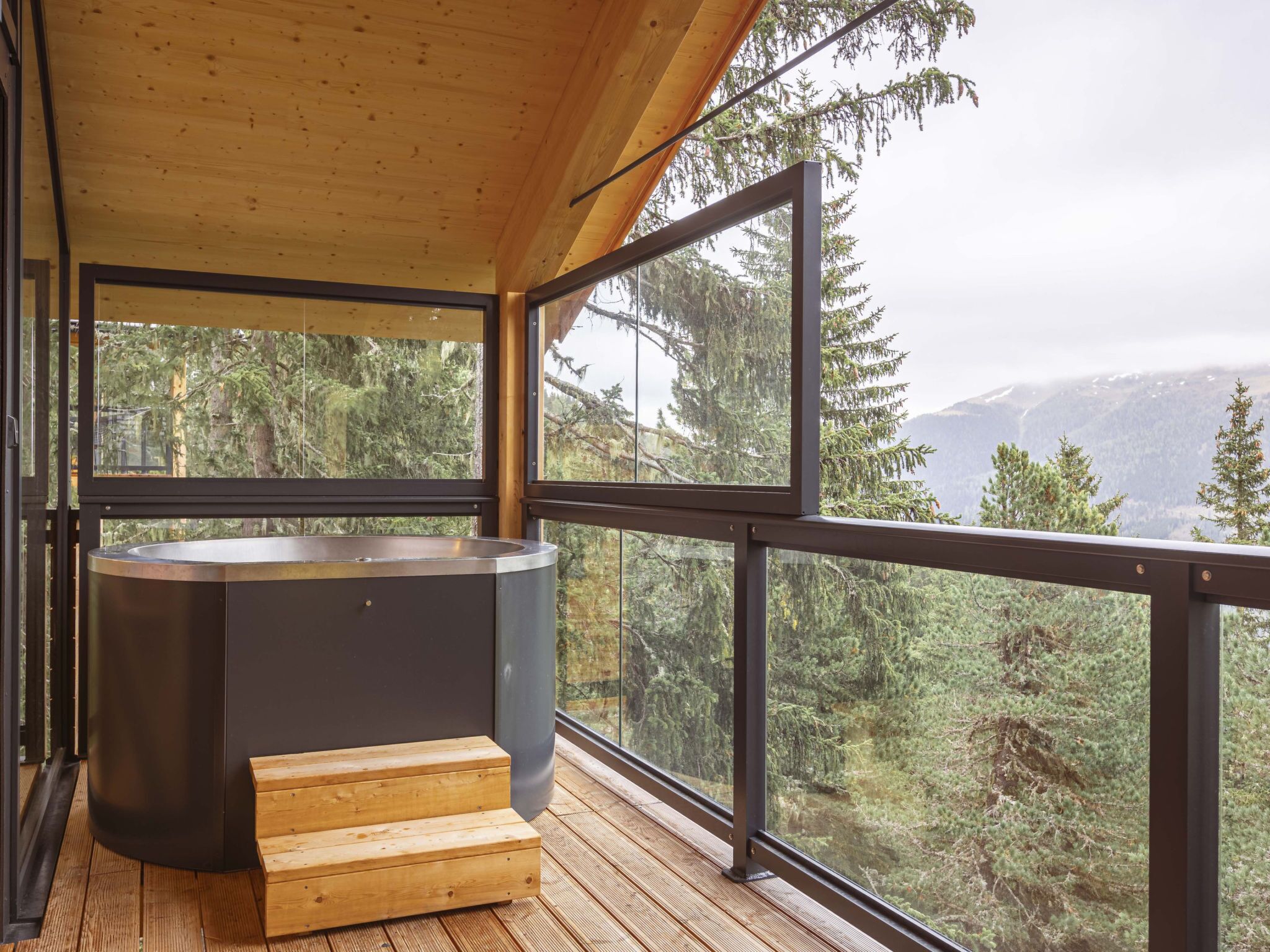 Une terrasse en bois dotée d’un jacuzzi et d’un escalier menant à un balcon offrant une vue sur une zone montagneuse boisée.