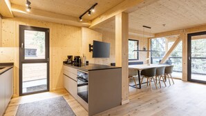 Une cuisine moderne avec des murs en bois et un îlot central, un coin repas avec une table et des chaises, et de grandes fenêtres offrant une vue sur l’extérieur.