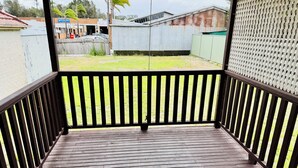Une terrasse en bois avec une rambarde noire, qui mène à une cour verdoyante, délimitée par une clôture en métal ondulé, et où l’on aperçoit un bâtiment au fond.