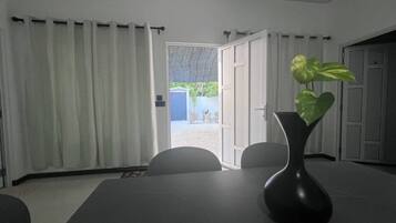 Ein Zimmer mit Tisch und Stühlen, eine schwarze Vase mit Pflanze sowie Blick auf einen Außenbereich mit Tür und blauer Mülltonne.