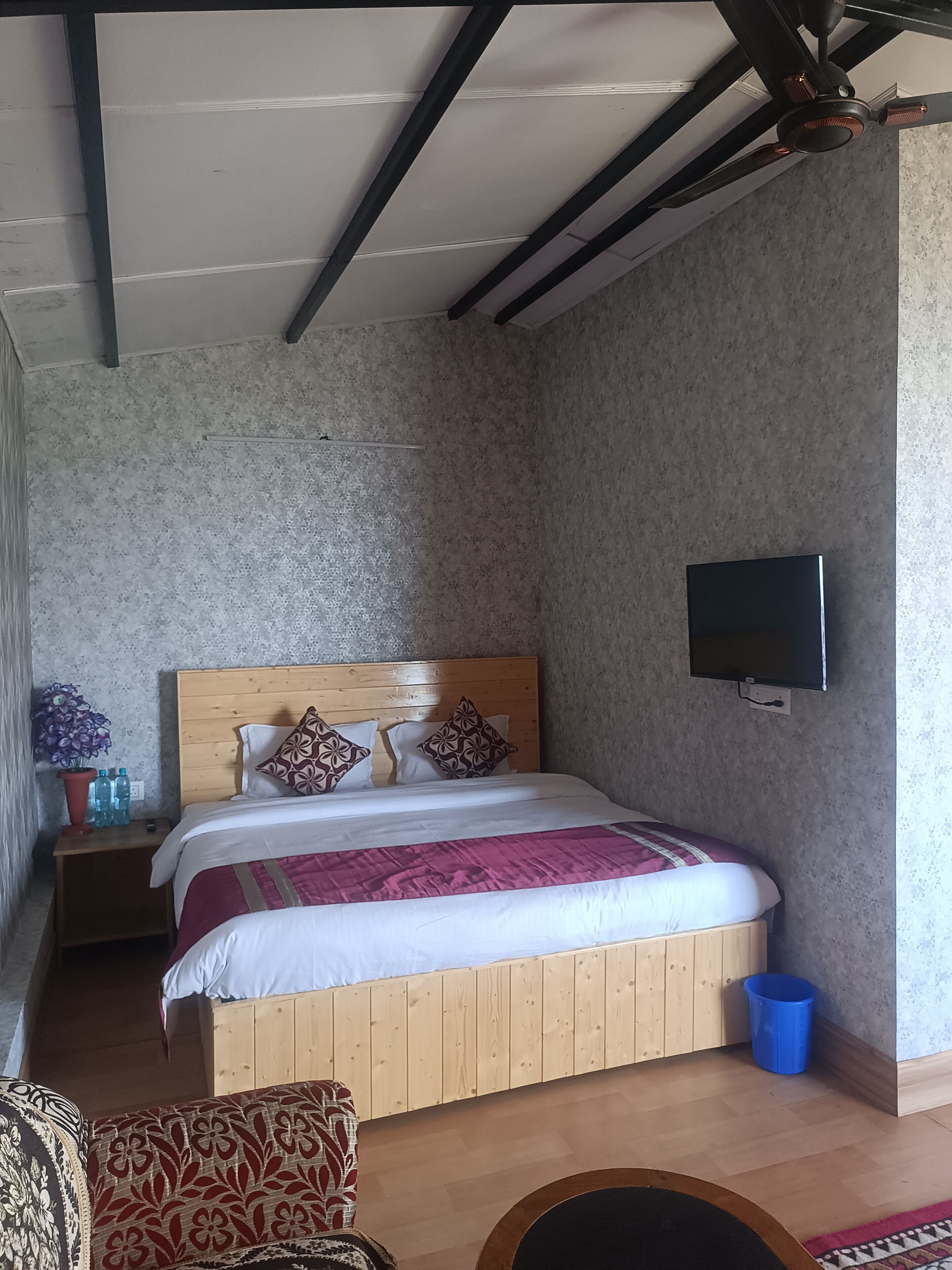 Un dormitorio con cama de madera, televisor montado en la pared, mesita de noche con botellas de agua y un cubo de basura azul.