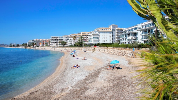 Ein Strand mit Gebäuden im Hintergrund, klares blaues Wasser und Menschen, die am Sand entspannen.