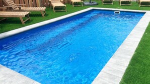 A rectangular swimming pool with a concrete edge, surrounded by artificial grass and wooden lounge chairs.