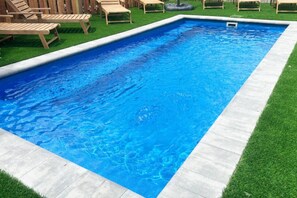 Une piscine rectangulaire bordée de béton, entourée de gazon synthétique et de chaises longues en bois.