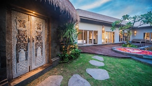 A modern house with a thatched roof, a stone wall with intricate carvings, and a well-maintained garden with stepping stones. - Luxury-3BedRoom-Sang Villa-Central Sanur (Bali)