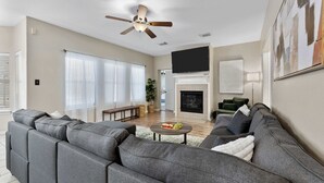 A modern living room with a sectional sofa, a flat-screen TV mounted above the fireplace, and a ceiling fan. - Luxurious Home with Great Pool Area (Willis)