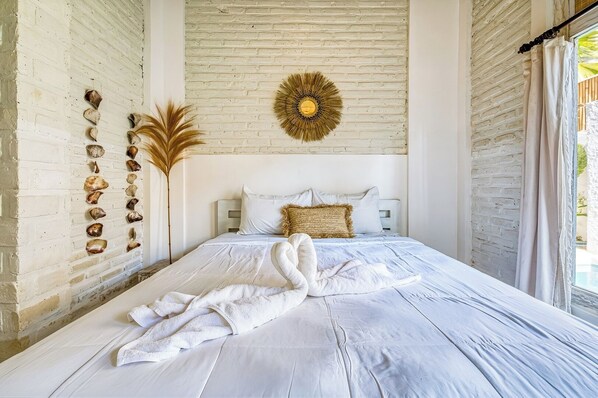 A bedroom with a white brick wall, a bed with white bedding, a decorative sunburst wall art, and a collection of seashells on the wall.