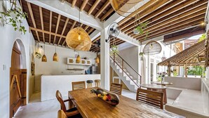 Un comedor espacioso con una mesa de madera dispuesta para cuatro personas, rodeada de sillas de madera. Se coloca un frutero en la mesa, y la habitación cuenta con vigas de madera a la vista y lámparas colgantes.