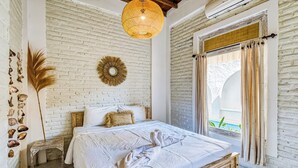 Un dormitorio con pared de ladrillo blanco, cama con ropa de cama blanca, lámpara colgante y vista al exterior.
