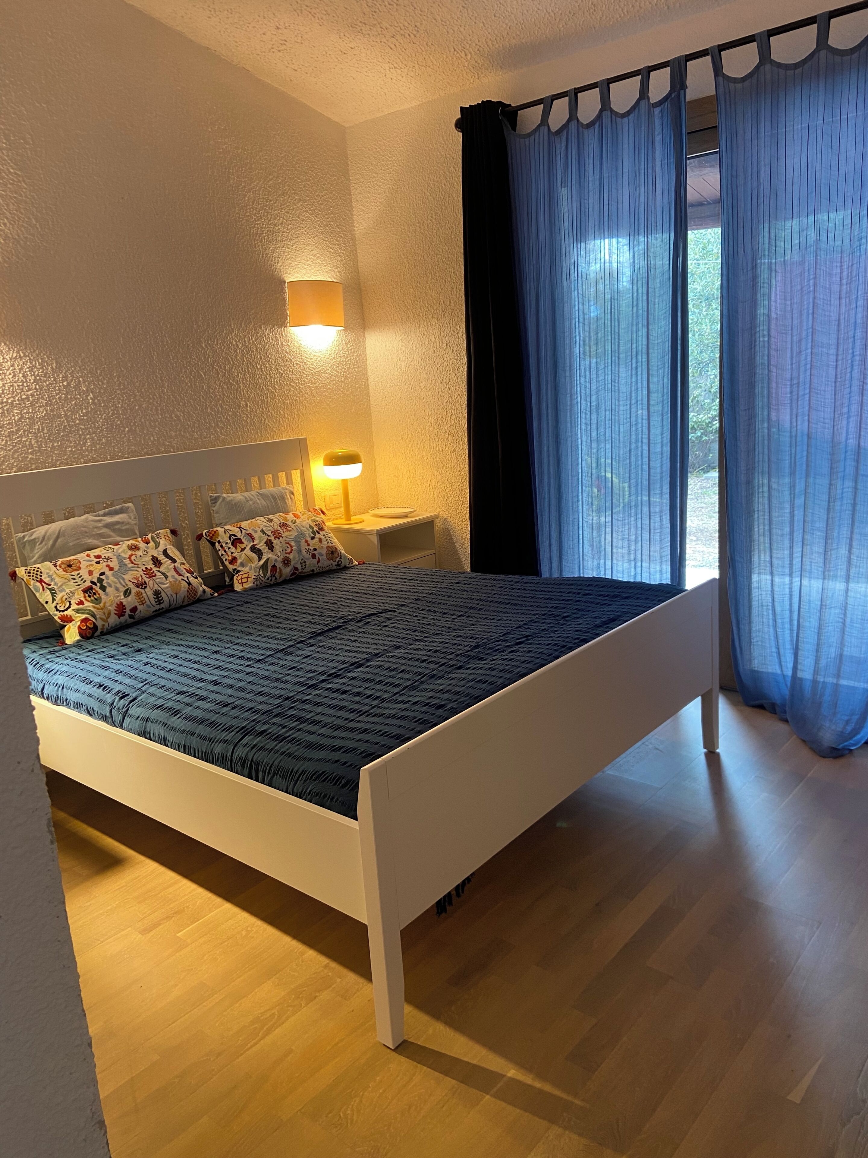 Un lit avec un cadre blanc, une couvre-lit bleue et deux oreillers à motifs floraux. On y trouve une lampe de chevet avec un abat-jour jaune, une petite table de chevet blanche et une fenêtre avec des rideaux bleus.