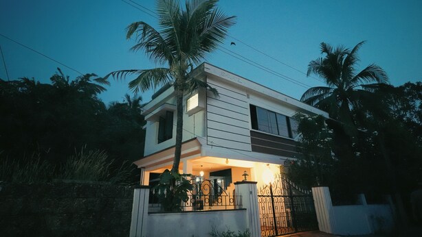 Una moderna casa a due piani con un balcone, circondata da palme e un ingresso con cancello.