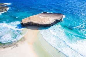 An aerial view of a beach with a large rock formation, clear turquoise waters, and a sandy shoreline.