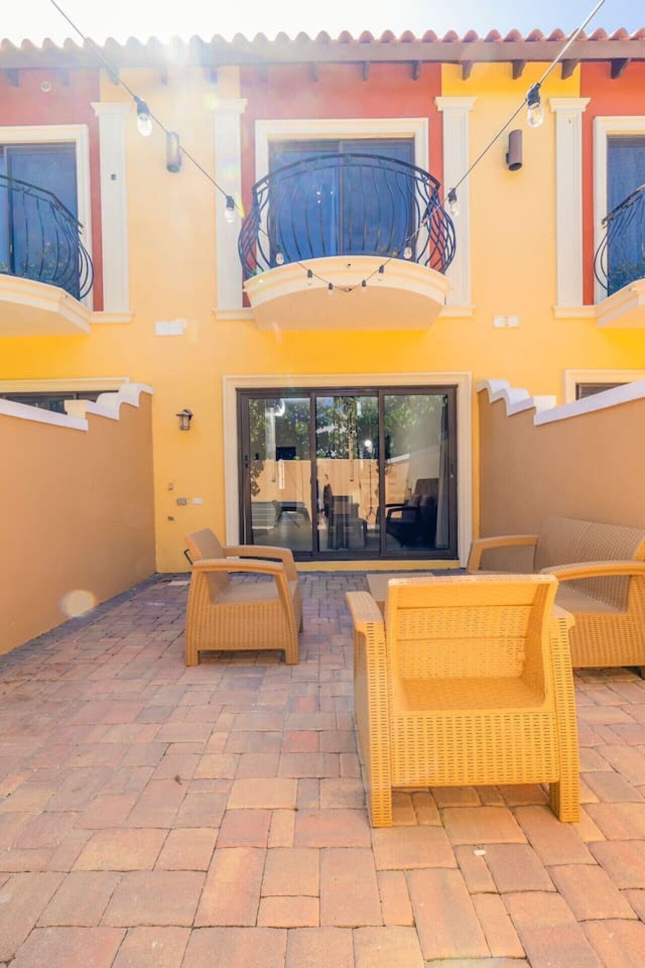 Un bâtiment à deux étages doté d’un balcon, d’un espace extérieur aménagé avec des meubles en osier et d’une entrée avec une porte vitrée.