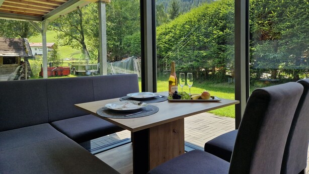 Een moderne eethoek buiten, met een houten tafel en beklede zitplaatsen, met uitzicht op een weelderig groonlandschap en een berg in de verte.