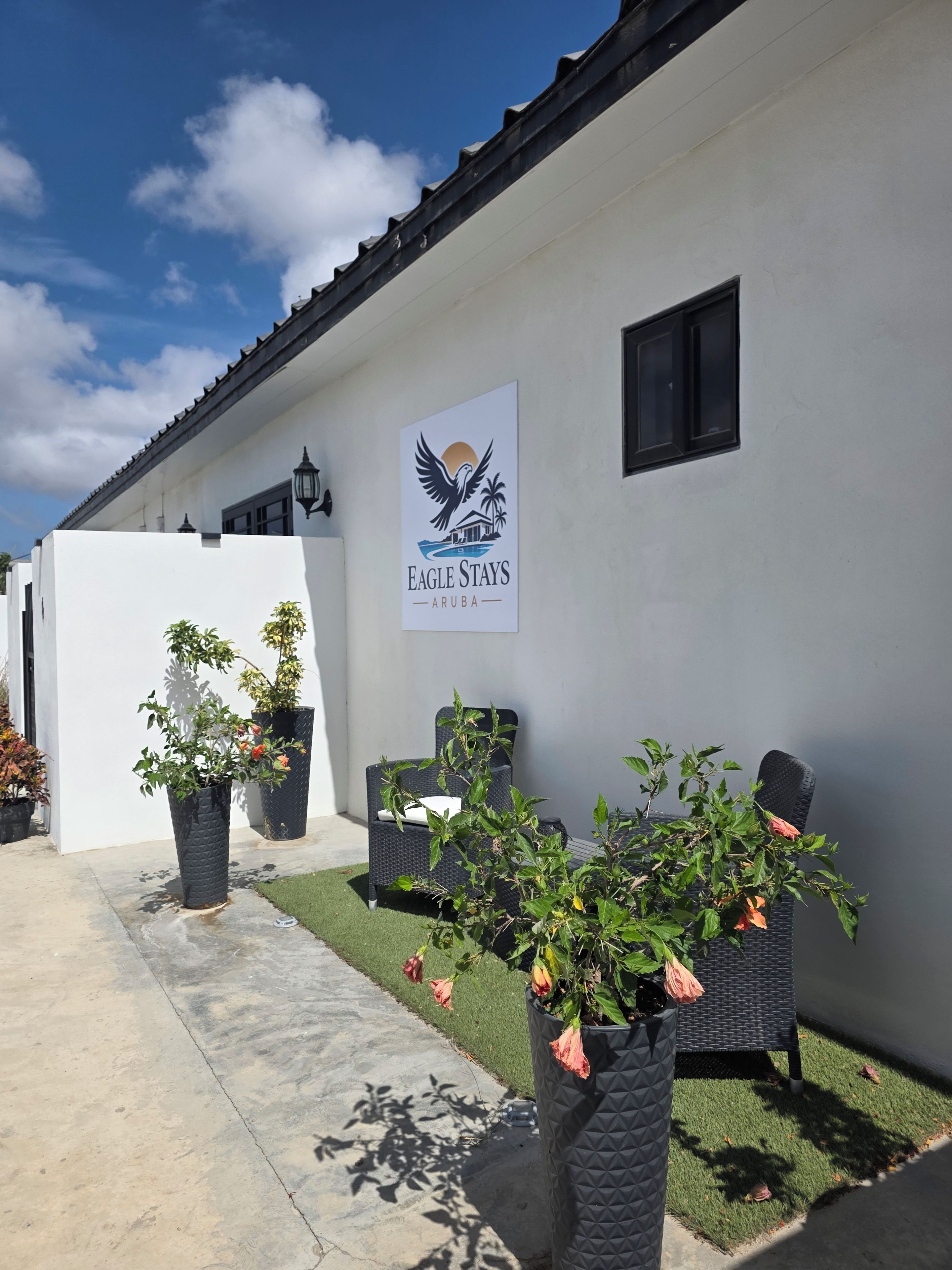 L’extérieur d’un hôtel, avec une enseigne indiquant « Eagle Stays Aruba », des places assises en plein air et des plantes en pot.
