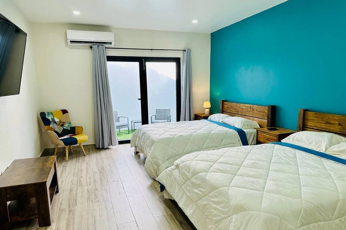 A hotel room with two beds, a chair, a small table, and a view of mountains through a large window.